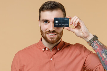 Happy smiling fascinating tatooed young brunet man 20s with short haircut wears apricot shirt hold in hand credit bank card cover close hiding eye isolated on pastel orange background studio portrait