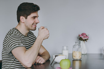 Young side view happy man in casual clothes striped t-shirt eat breakfast muesli cereals with milk fruit in bowl drink coffee cooking food in light kitchen at home alone Health diet lifestyle concept.