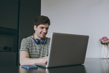 Young smiling happy satisfied fun man 20s wearing striped t-shirt headphones using laptop pc computer working online browsing sitting by table in light kitchen at home alone. People lifestyle concept