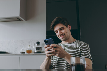 Young happy cheerful smiling man in striped t-shirt use mobile cell phone browsing chatting online with friends typing message sitting by table in light kitchen at home alone People lifestyle concept