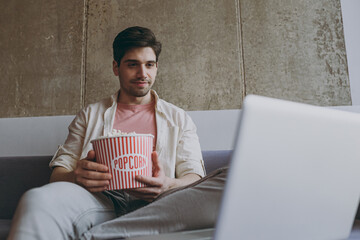 Young smiling happy man 20s wear casual clothes use laptop pc computer watch series hold takeaway popcorn bucket eat snack sit on grey sofa rest indoors at home on weekends People leisure concept.