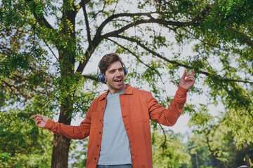 Bottom view fun young man in orange jacket blue t-shirt headphones walk listen music rest relax in spring green city park go down alley sunshine lawn outdoor on nature Urban lifestyle leisure concept