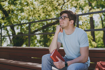 Lower young pensive student man in blue t-shirt eyeglasses prop up chin read book sit on bench walking rest relax in sunshine spring green city park outdoors on nature. Education high school concept