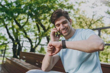 Smiling young happy man wear blue t-shirt sit on bench talk speak on mobile cell phone check time...