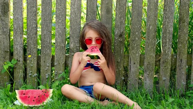 A cute girl eats a piece of watermelon with pleasure sitting on the grass near the fence in the yard. National Watermelon Day. Sweet juicy delicious refreshing watermelon