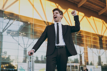 Bottom view happy young traveler brunet businessman man in black classic tie suit stand outside at international airport terminal with suitcase do winner gesture Air flight business lifestyle concept