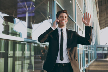 Bottom view young traveler businessman man in black suit stand outside at international airport terminal talk on mobile cell phone book taxi order hotel waving hand Air flight business trip concept.
