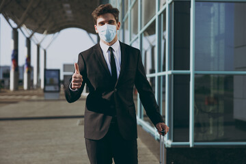 Satisfied young traveler businessman man in black dinner suit sterile facial mask walk go outside at international airport terminal with suitcase valise show thumb up Air flight business trip concept.