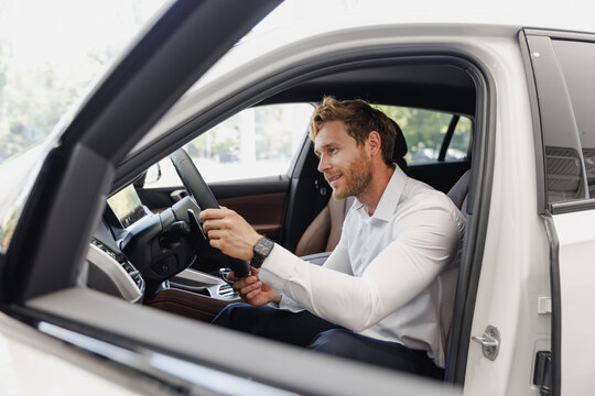 Adult man customer male buyer businessman client wears classic suit sit in car salon chooses auto wants to buy new automobile in showroom vehicle dealership store motor show indoor. Car sales concept.