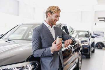 Man customer buyer client in suit choose auto drink tea hold cup coffee look at time smart watch want buy new automobile in car showroom vehicle salon dealership store motor show indoor Sales concept