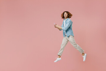 Fototapeta na wymiar Full length side view young excited happy man with long curly hair wear blue shirt white t-shirt using hold in hand mobile cell phone jump high isolated on pastel plain pink color background studio