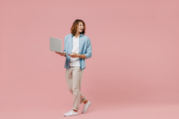 Full length young minded happy man with long curly hair in blue shirt white t-shirt hold use work on laptop pc computer look aside isolated on pastel plain pink color wall background studio portrait