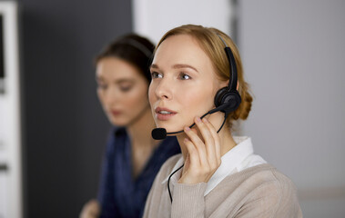 Friendly businesswoman talking by headset while sitting in office. Call center and diverse people...