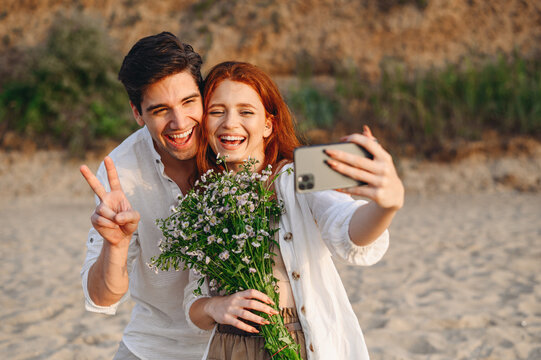 Young couple family man woman in white clothes rest together show bouquet v-sign do selfie shot mobile cell phone post photo social network at sunrise over sea sand beach outside seaside in summer day