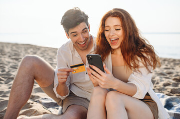 Young couple two man woman in white clothes sit on sand using mobile phone credit card shopping online order delivery booking tour rest together at sunrise over sea beach outside seaside in summer day