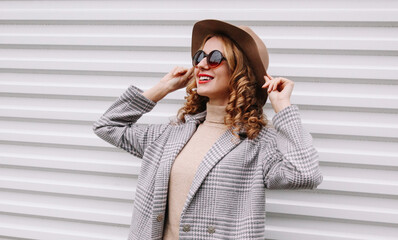 Fashionable portrait of happy smiling young woman model with curly hair looking away wearing a checkered coat, round hat on city street on white background