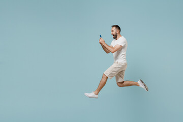 Full length side view young overjoyed happy man 20s wearing casual white t-shirt jump high using mobile cell phone chatting online isolated on plain pastel light blue color background studio portrait