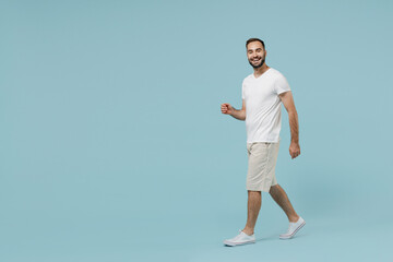 Fototapeta na wymiar Full length side view young friendly fun smiling happy cheerful man 20s in casual white t-shirt walk go strolling looking camera isolated on plain pastel light blue color background studio portrait