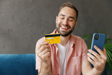 Young cool man 20s in casual clothes use mobile cell phone hold credit bank card do online shopping order delivery book tour sitting on blue sofa at home flat indoors rest relax on weekends free time