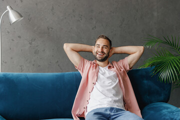 Young smiling happy fun dreeamful relaxed man 20s wearing casual clothes hold hands behind neck sitting on blue sofa at home flat indoors rest on weekends free time. People lounge lifestyle concept