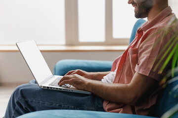 Cropped close up side view young smiling man in casual clothes hold use work on laptop pc computer look aside sitting on blue sofa at home flat indoors rest relax on weekends. People lounge concept