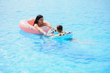 Happy family, young active mother and adorable curly little baby having fun in a swimming pool, child learning to swim in an inflatable toy ring, enjoying summer vacation at a tropical resort