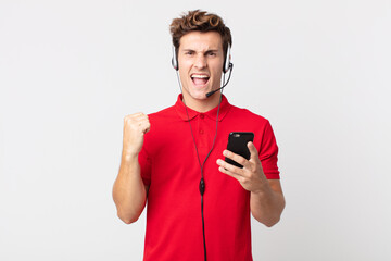 young handsome man shouting aggressively with an angry expression with a smartphone and headset