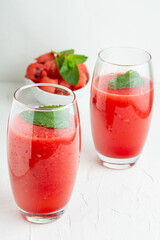 Aerial view of two glasses with watermelon juice, mint, and bowl with pieces of watermelon, selective focus, on table and white background, in vertical, with copy space