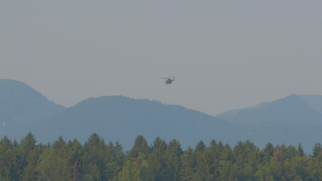 Slow motion view of military helicopter flying over forest. Alps mountains in the distance. Chopper fly away. Ground view of army helicopter in the air