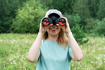 happy Young blonde woman bird watcher in cap and blue t-shirt looking through binoculars at cloudy...