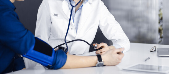 Unknown woman doctor is checking her patient's blood pressure while sitting together in clinic. Medicine concept during Coronavirus pandemic. Covid 2019