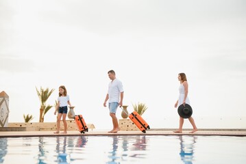 young family with suitcases walking to hotel building with beautiful swimming pool. traveling and relaxing time concept.