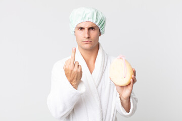 young handsome man feeling angry, annoyed, rebellious and aggressive with bathrobe, shower cap and...