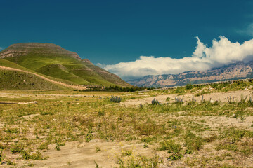 Fototapeta na wymiar Toned mountain landscape view. Summer cloudy day in valley. Panorama with high hills, light brown earth, green grass and background of blue sky with white clouds.