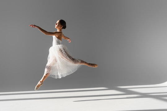 Side view of professional ballerina jumping on grey background with sunlight