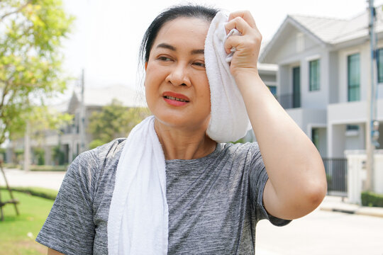 Asian Female Runner Tired Of Exercise Running In The Morning At The Neighborhood. Woman Using Towel To Wipe The Sweat After Workout 