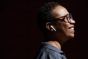 Close-up of African young woman in eyeglasses listening to music in wireless headphones and smiling isolated on black background