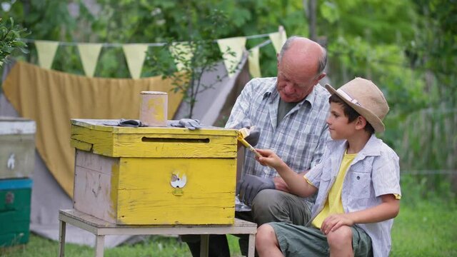cute male child helping his grandfather beekeeper paint beehive with yellow paint, smile, look at camera and point up while sitting background of green trees