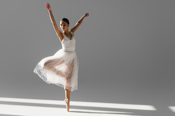 Brunette ballerina in white skirt dancing with closed eyes on grey background with sunlight - obrazy, fototapety, plakaty