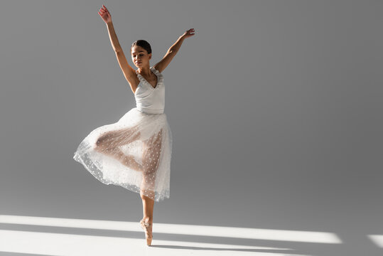 Brunette ballerina in white skirt dancing with closed eyes on grey background with sunlight