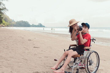 Fototapeta na wymiar Happy disabled teenage boy smile face on wheelchair holding a camera with mother, Activity outdoors with family on the beach background, People having fun and diverse people concept.