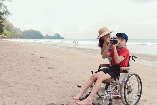 Happy disabled teenage boy smile face on wheelchair holding a camera with mother, Activity outdoors with family on the beach background, People having fun and diverse people concept.