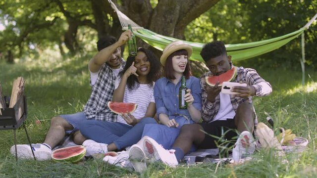 Young multiracial people using modern smartphone for taking selfie at green garden. Four cheerful friends having fun during picnic in nature, eating watermelon and gesturing.