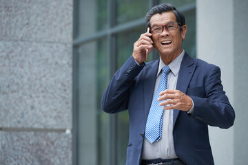 Portrait of positive aged businessman making phone call to client or business partner when standing at entrance of office building