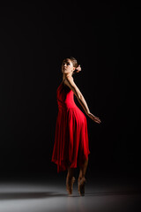 Naklejka premium Young ballerina in red dress and pointe shoes dancing on black background