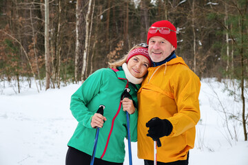Smiling senior couple walking with nordic walking poles in snowy winter forest. Elderly wife and husband doing healthy exercise outdoors. Active lifestyle after retirement concept.