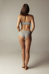 Naklejka premium Portrait of young beautiful slim woman in lingerie posing isolated over gray studio background. Natural beauty concept. Back view
