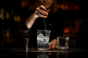 hand of female bartender holds spoon and stirred cold cocktail in crystal mixing cup.