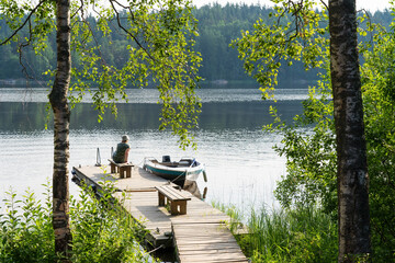 The woman sits on a wooden pier on a large lake. There is a moored motor boat. Trees are in the...