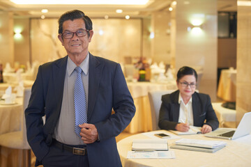 Portrait of smiling confident senior entrepreneur in suit standing at restaurant table, his colleague working with documents in background
